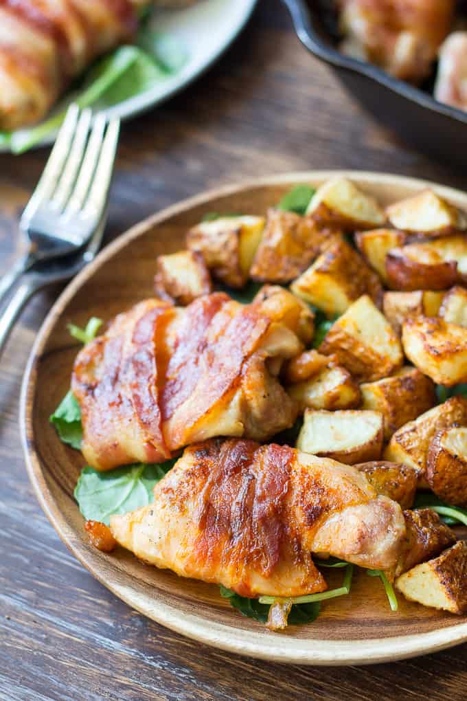 bacon wrapped chicken with crispy potatoes over spinach on a wooden plate