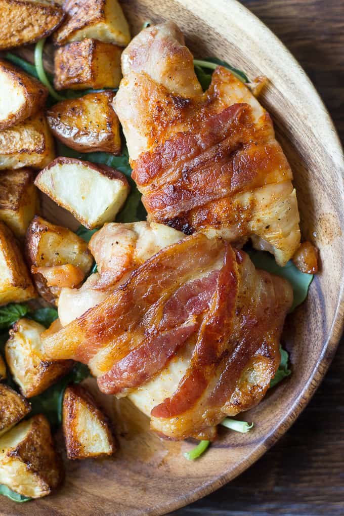 chicken thighs wrapped in bacon aside crispy potatoes over spinach on a plate