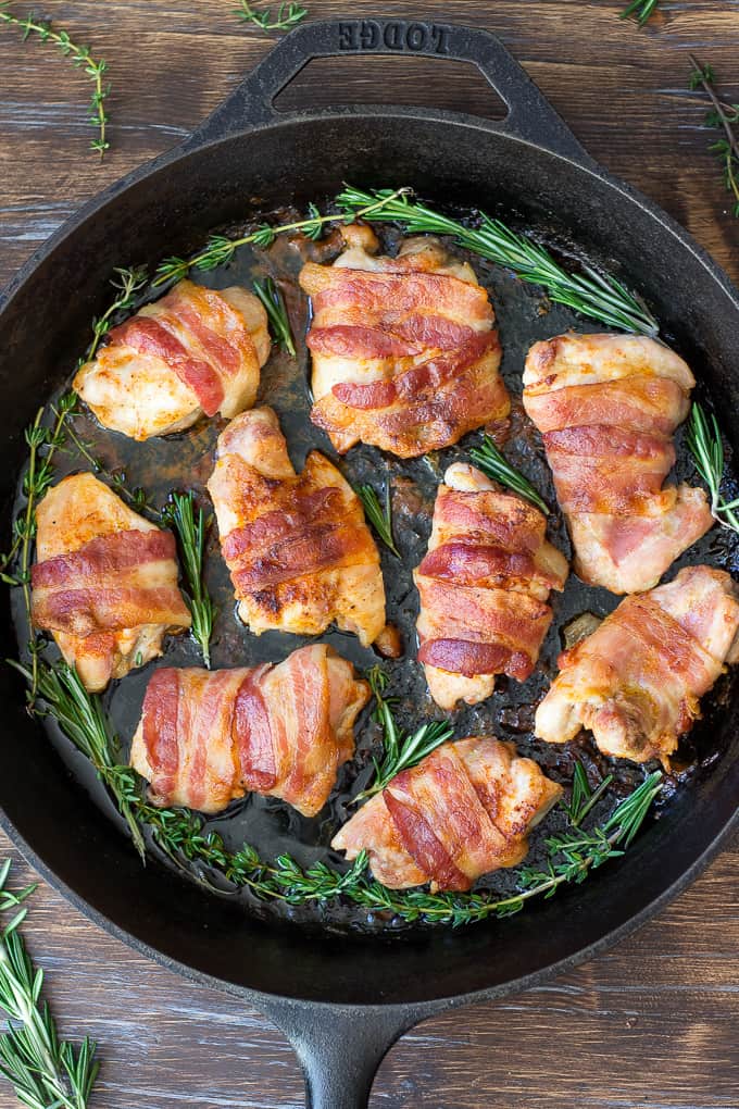 bacon wrapped chicken thighs in a cast iron skillet with rosemary and thyme sprigs