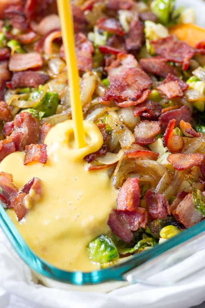 Scrambled eggs being poured over a dish of bacon, onions, and brussels sprouts