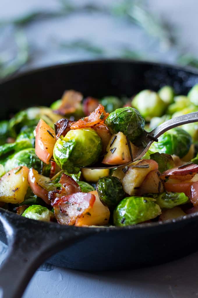 a spoon lifting a scoop of Brussels sprouts with apples, bacon, and rosemary from a cast iron skillet