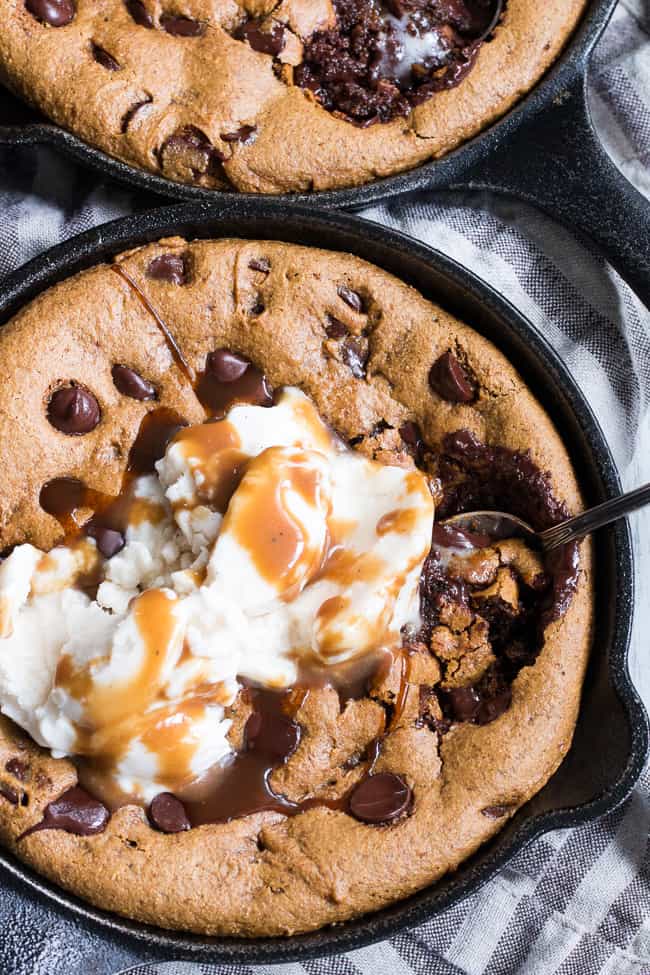 close up view of a chocolate chip cookie in a black skillet with white ice cream and caramel drizzle, and a spoon digging into the cookie