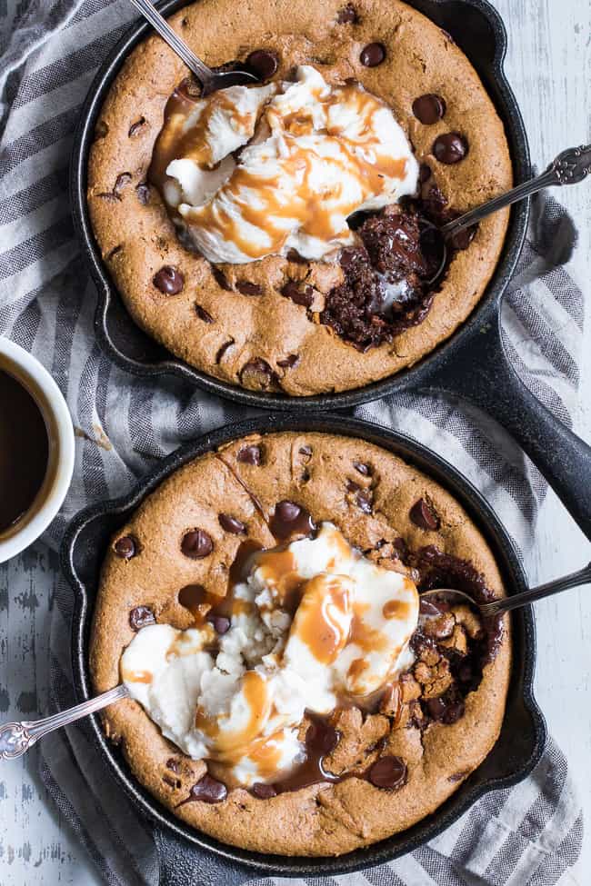 two black cast iron skillets with chocolate chip cookies topped with white ice cream scoops and caramel drizzled over top