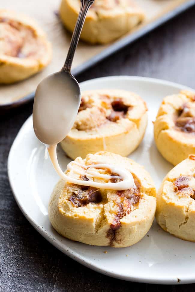 a spoon drizzling white frosting over a plate of cinnamon rolls