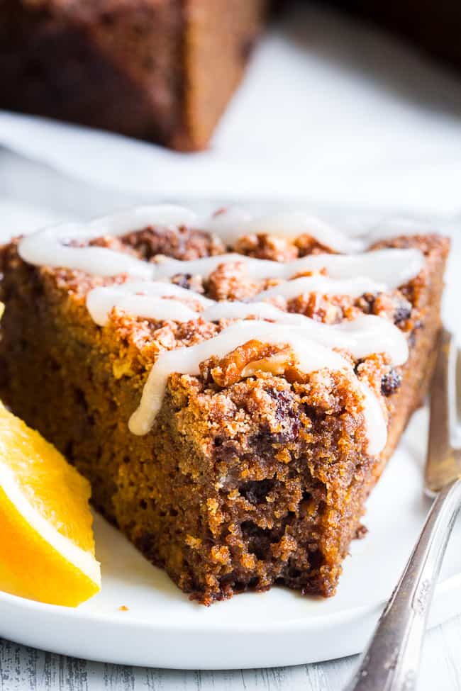 a triangle slice of brown coffee cake with white icing drizzled over top on a white plate with a slice of orange