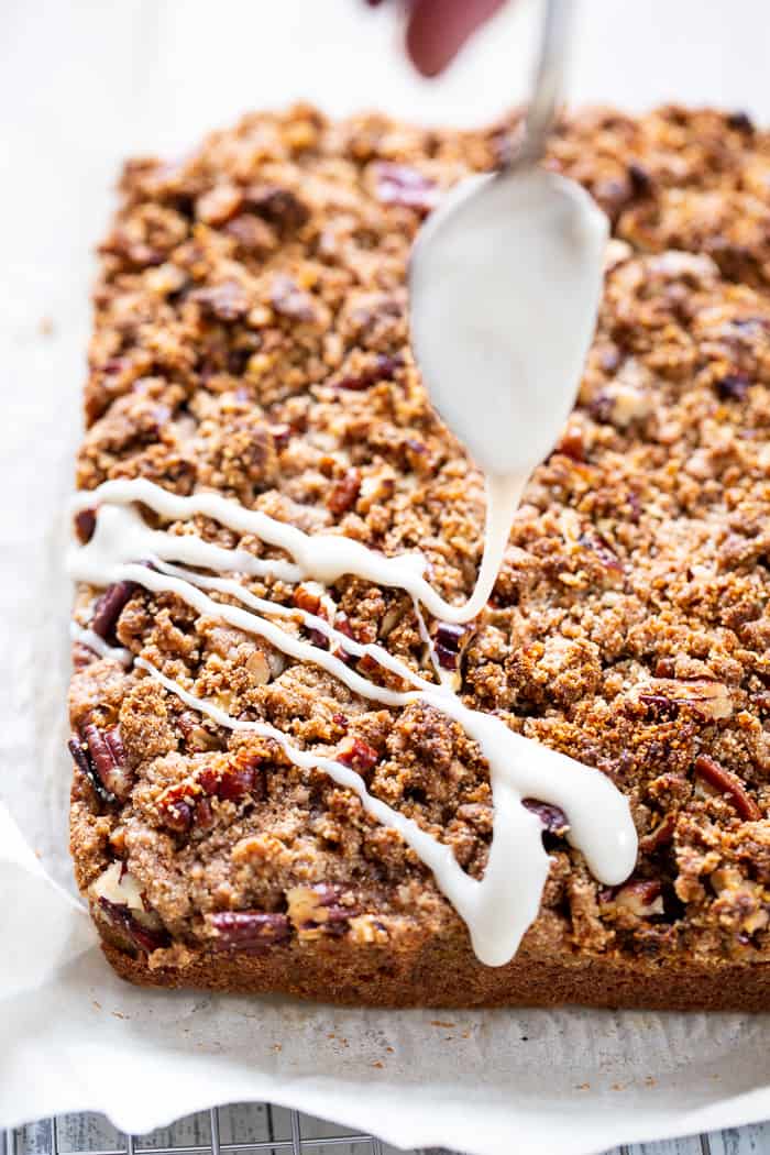 white frosting being drizzled with a spoon over the corner of a brown crumble topped cake slab