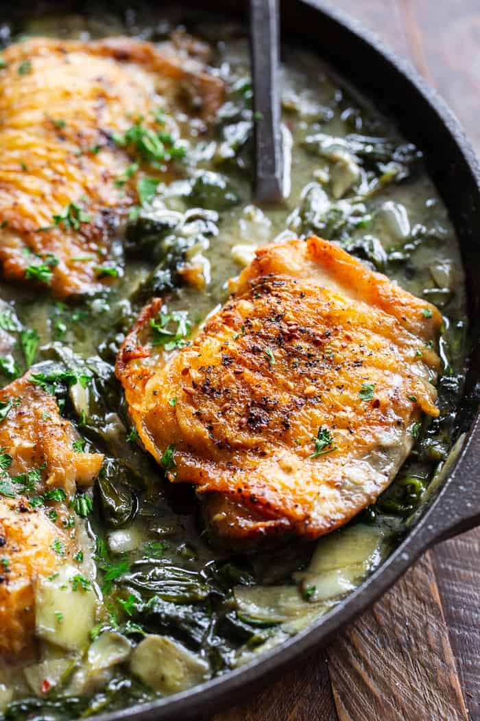 closeup shot of a cooked chicken thigh over spinach and artichokes in a cast iron skillet