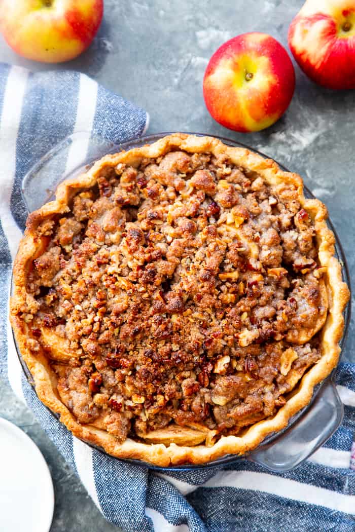 a baked pie with a browned crumble topping with whole red apples to the side and a blue and white striped napkin